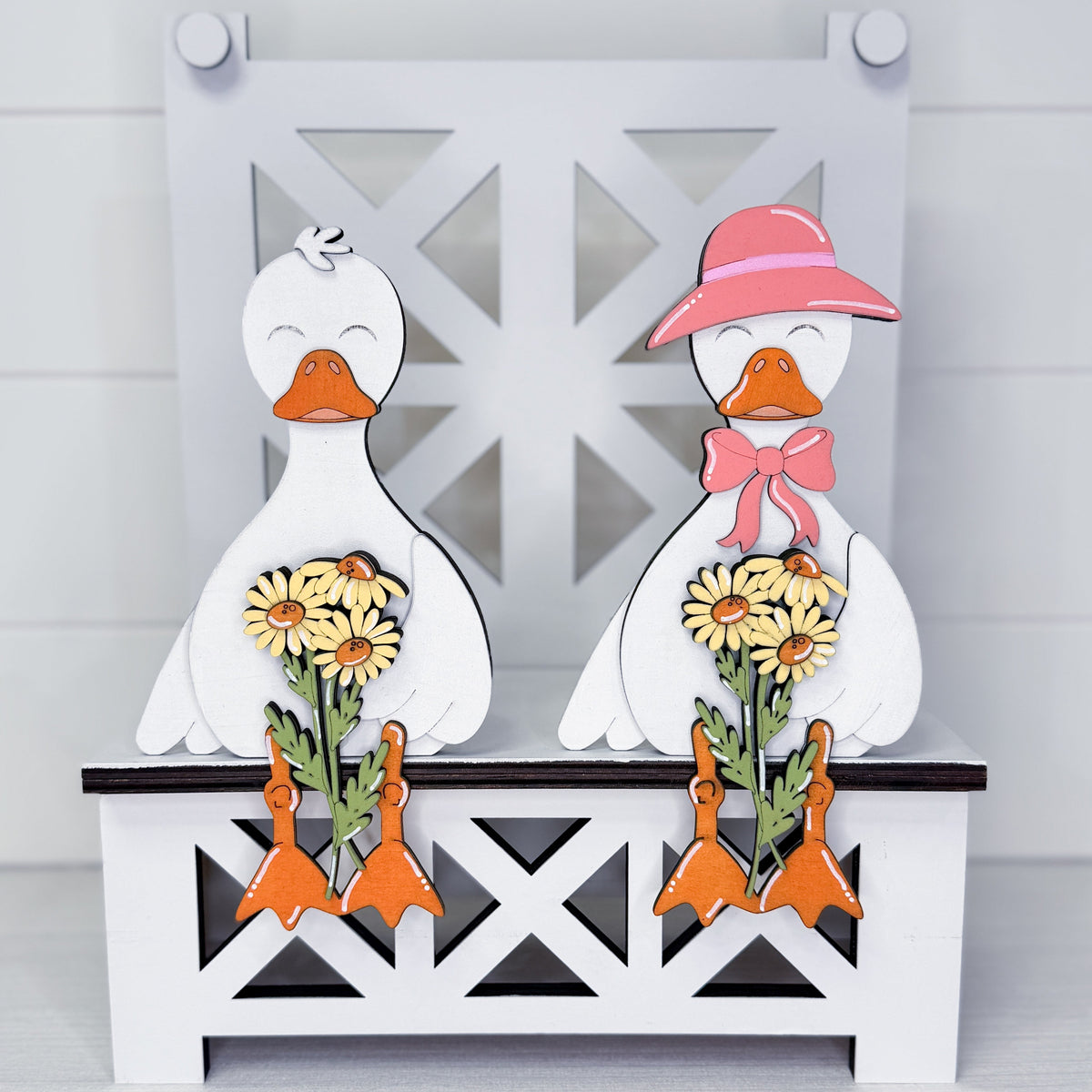A pair of white decorative ducks standing on a white bench. The left duck has an orange beak and holds yellow flowers with orange feet.