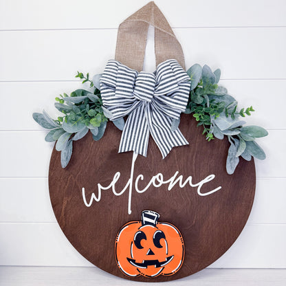 Decorative Halloween sign with a pumpkin and 'welcome' text on a wooden board with greenery and a bow.