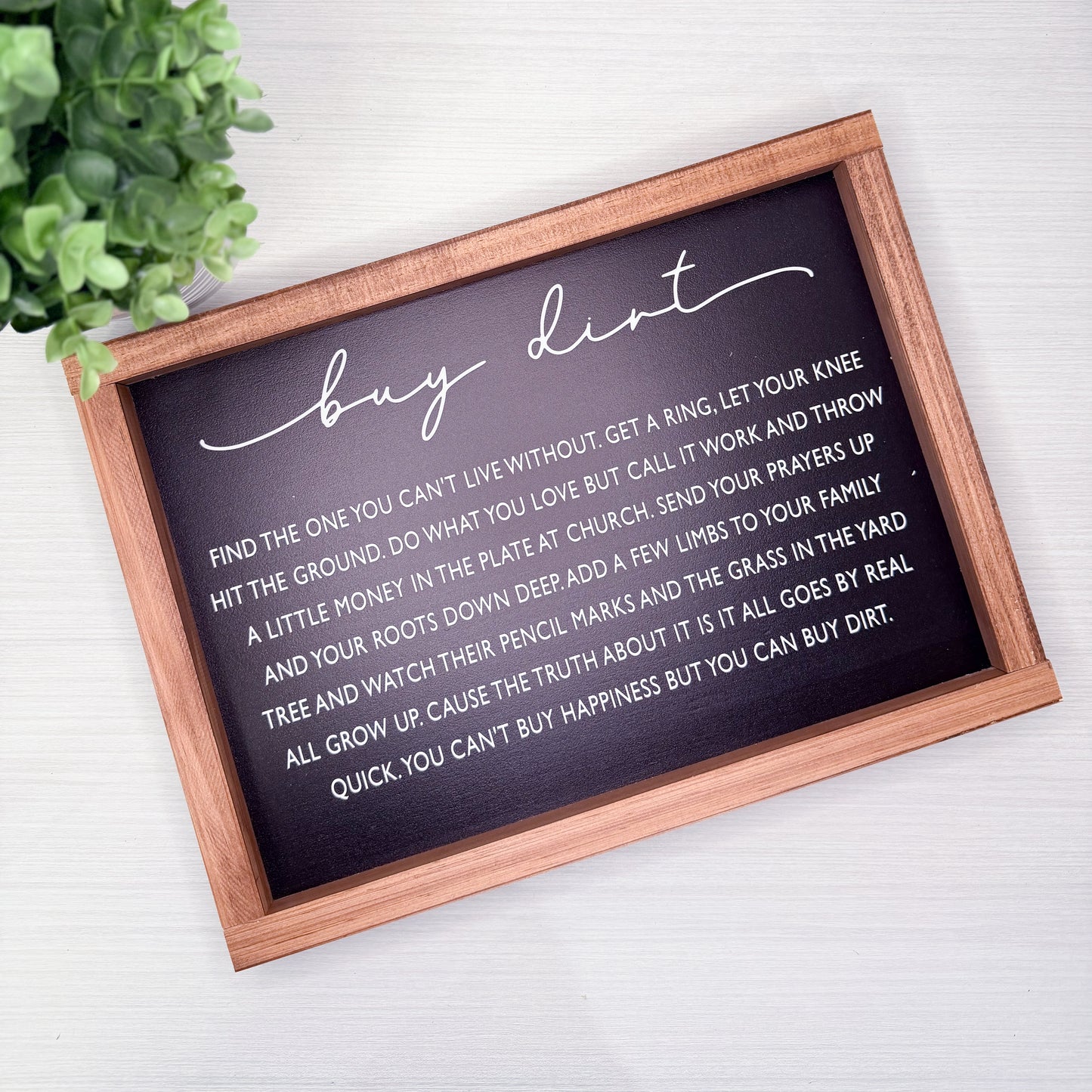 Decorative sign with text on a white surface with a plant in the corner