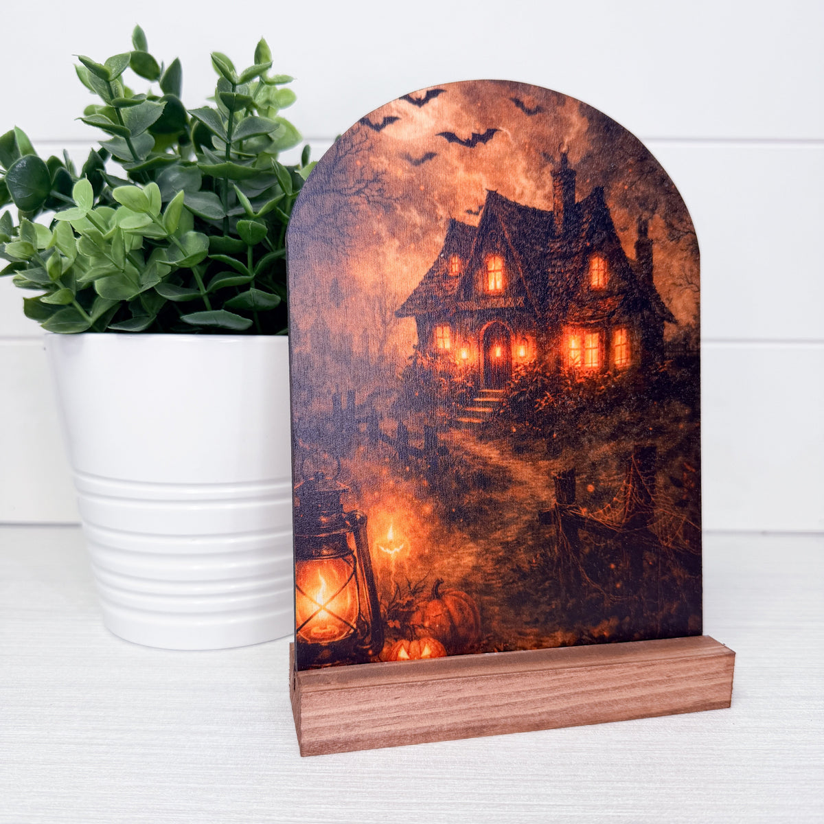 A Halloween-themed tabletop sign with a wooden base, featuring an arched panel printed with a spooky haunted house scene under a full moon, displayed next to a potted green plant on a white surface.