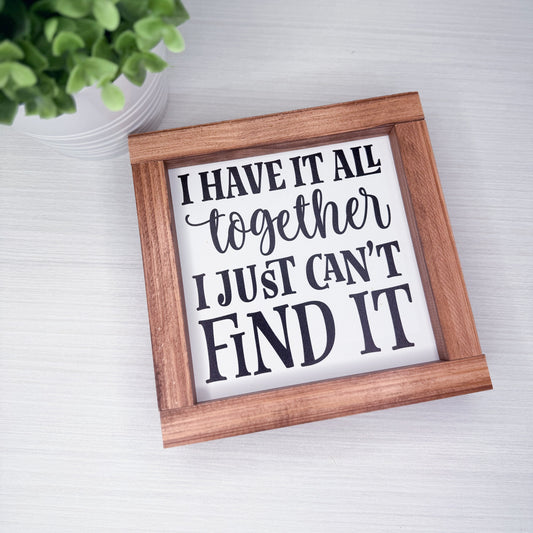 Wooden-framed sign with humorous text on a white surface with a plant in the corner.