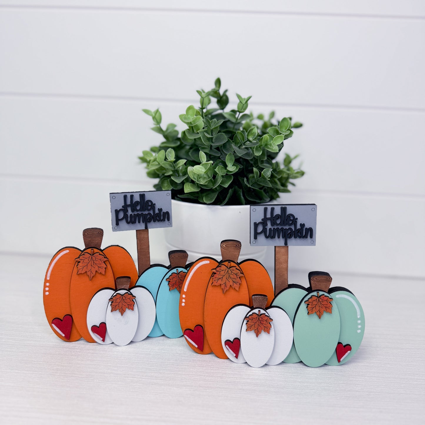 Two sets of decorative wooden autumn pumpkin signs with small 'Hello Pumpkin' labels, displayed in front of a potted green plant on a white surface against a white paneled background.
