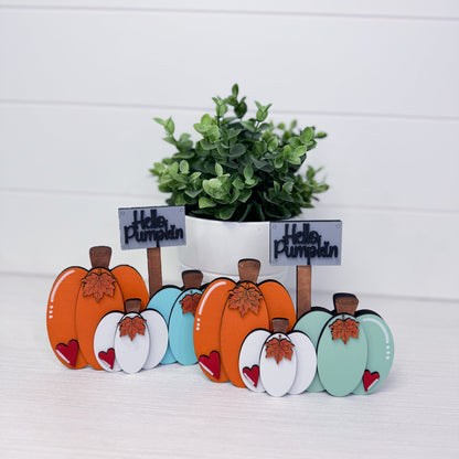 Two sets of decorative wooden autumn pumpkin signs with small 'Hello Pumpkin' labels, displayed in front of a potted green plant on a white surface against a white paneled background.