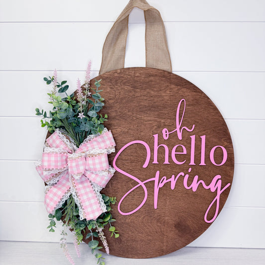 Wooden circular sign with 'oh hello Spring' text, pink bow, and greenery on a white background
