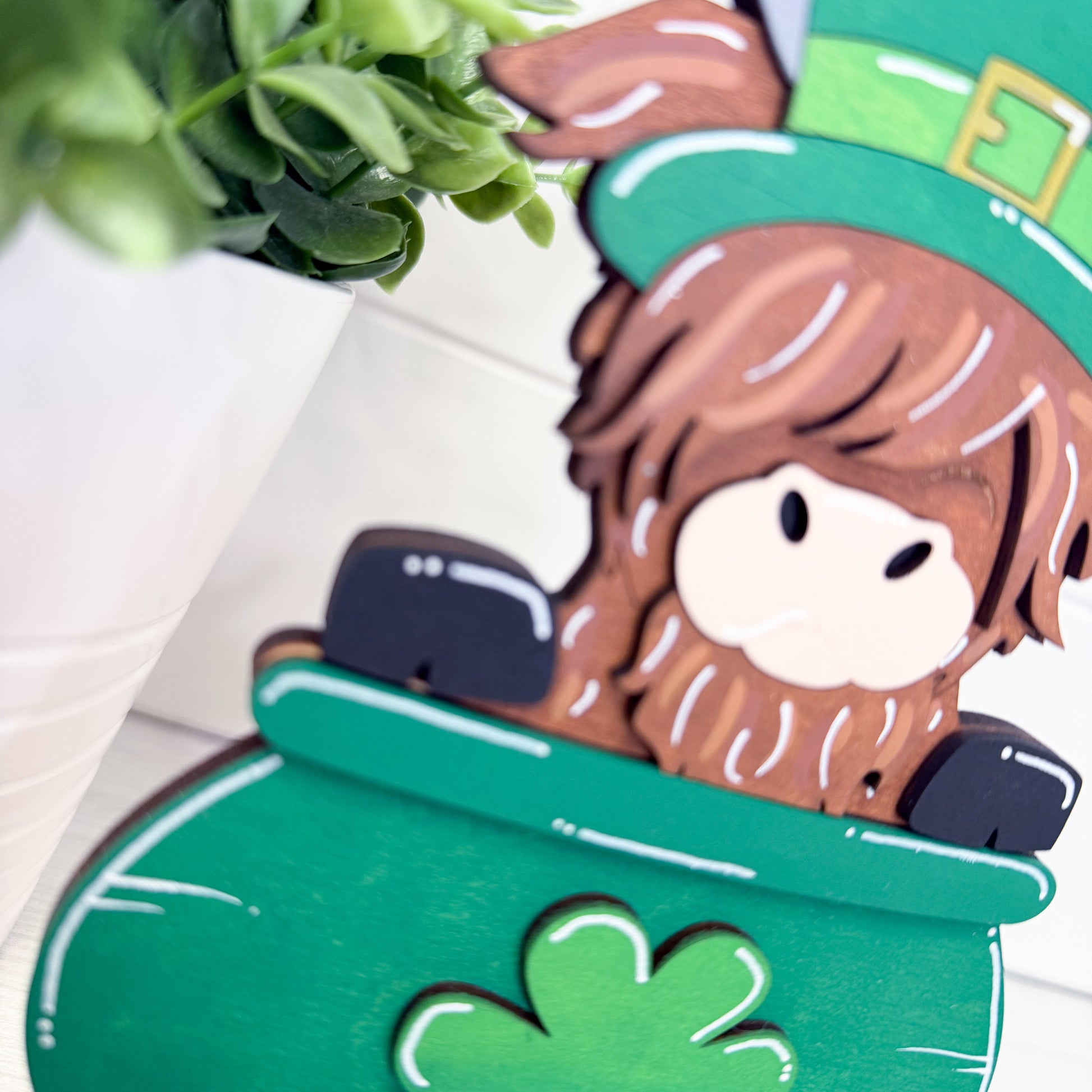 highland cow with brown hair and a green hat sitting in a pot of gold with a shamrock.