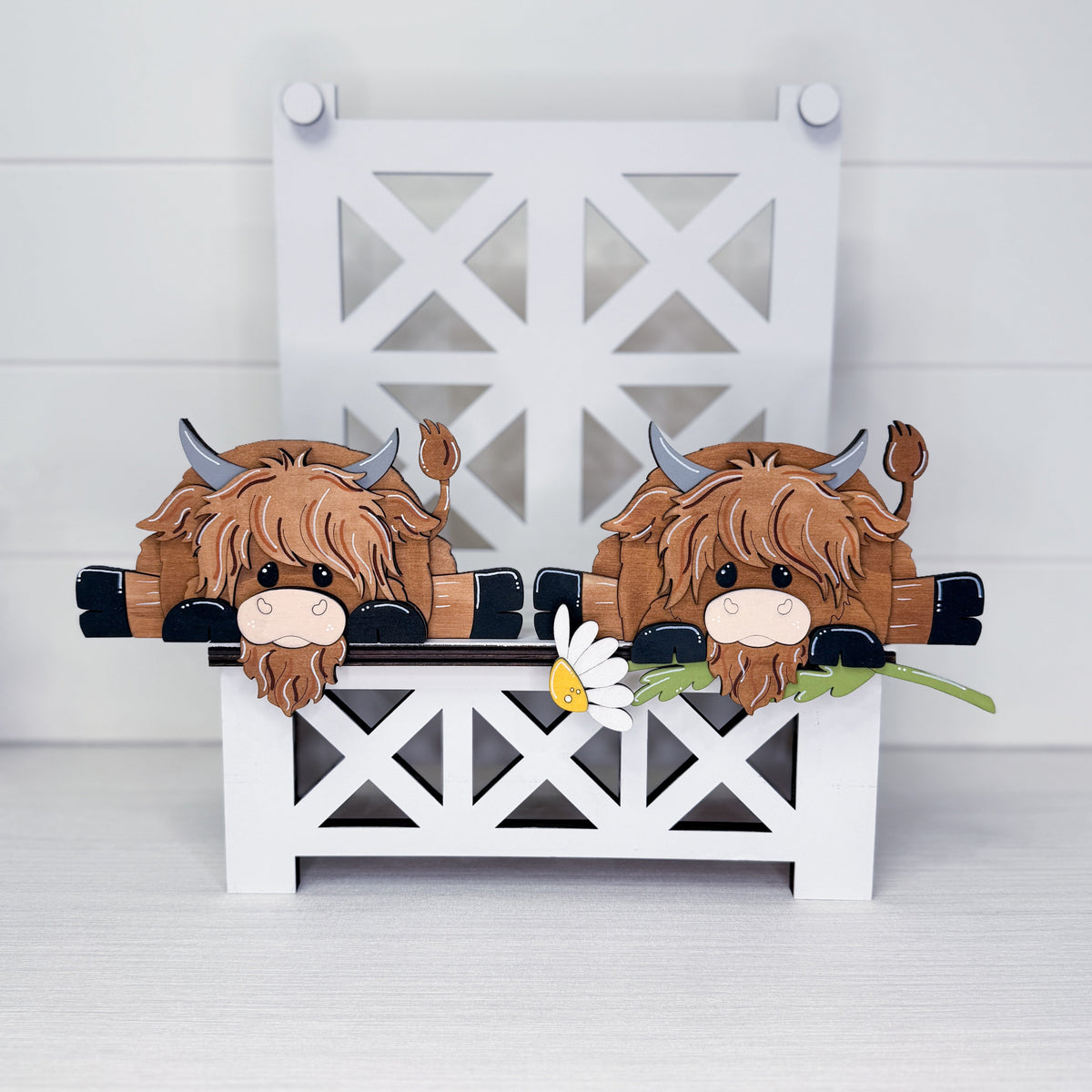 A decorative wooden Highland cow bench scene with two brown shaggy cows peeking over a white bench, a small daisy in front, and a white lattice bench backdrop on a light gray surface.