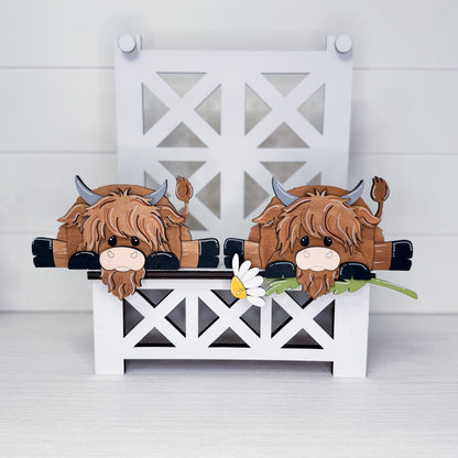A decorative wooden Highland cow bench scene with two brown shaggy cows peeking over a white bench, a small daisy in front, and a white lattice bench backdrop on a light gray surface.