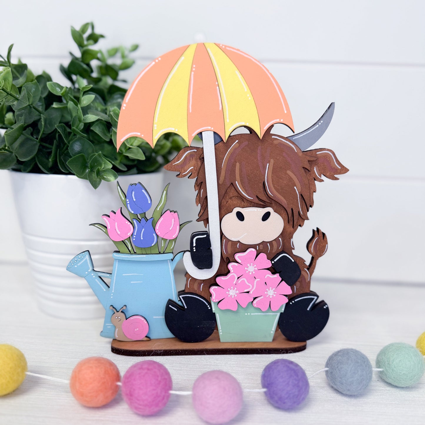 Decorative figurine of a cow holding an umbrella with flowers and a watering can, surrounded by plants and colorful balls.
