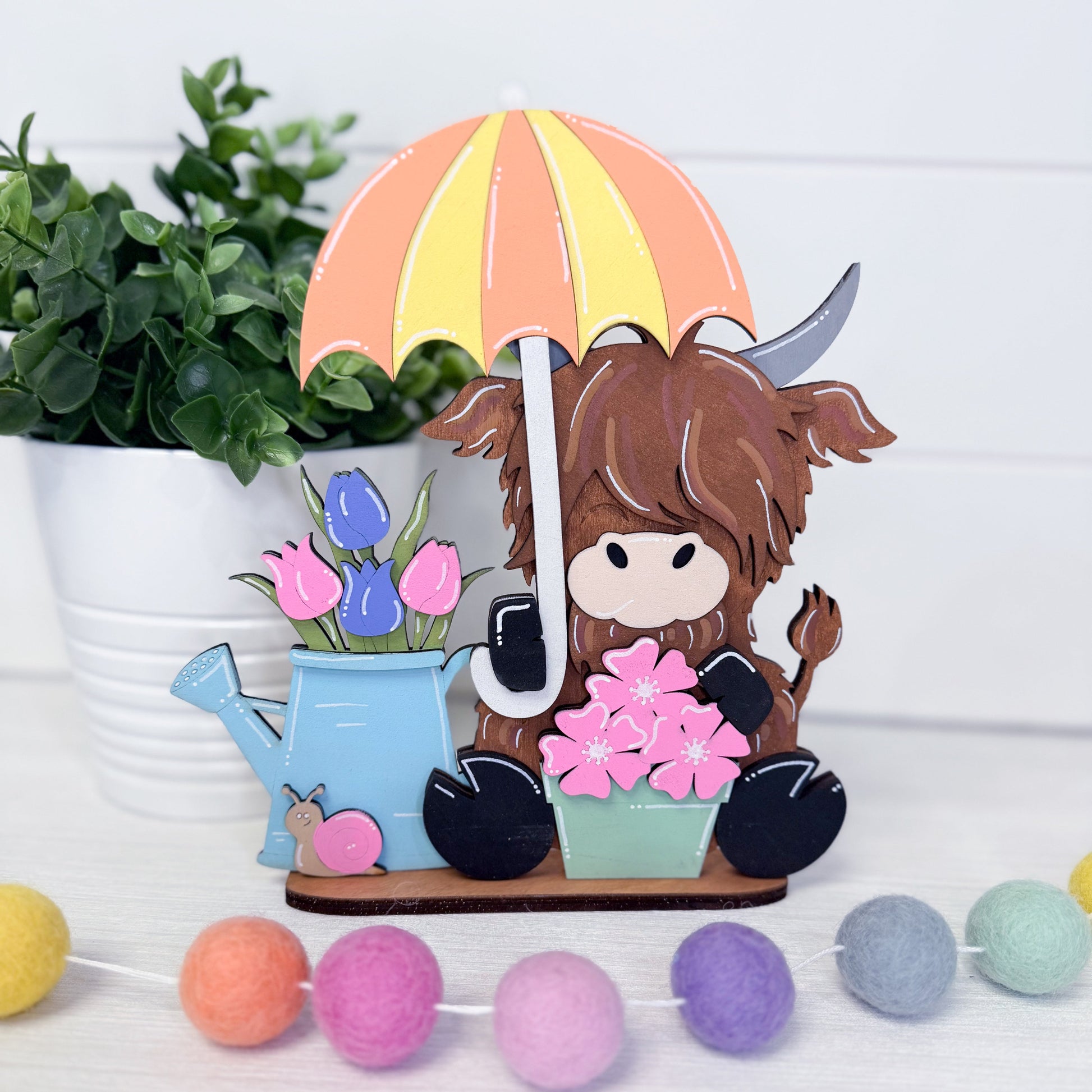 Decorative figurine of a cow holding an umbrella with flowers and a watering can, surrounded by plants and colorful balls.