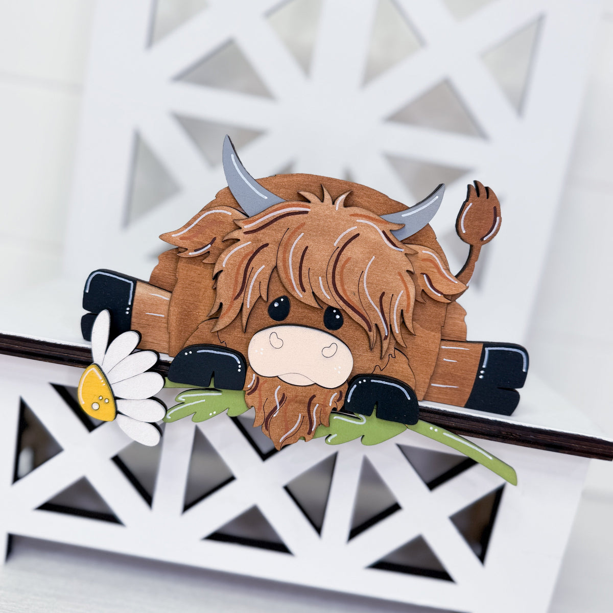 A wooden Highland cow figurine with brown shaggy hair, gray horns, and a small white daisy, displayed on a white geometric wooden stand.