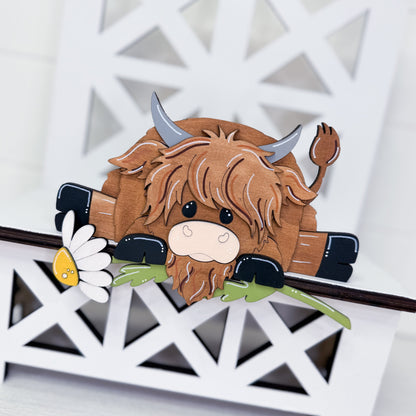 A wooden Highland cow figurine with brown shaggy hair, gray horns, and a small white daisy, displayed on a white geometric wooden stand.