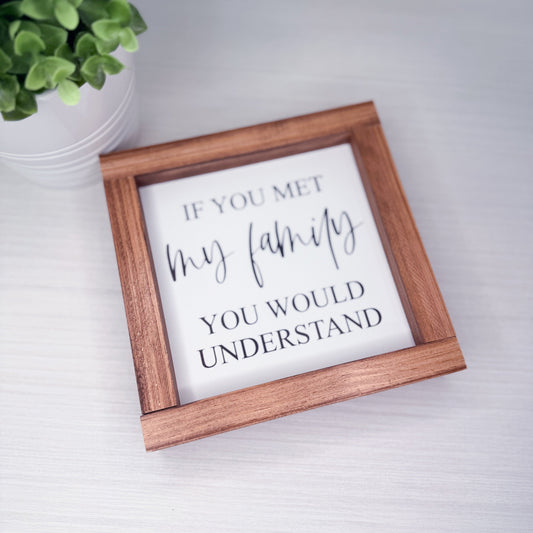 Wooden-framed sign with 'If you met my family you would understand' text on a white surface with a plant in the corner.