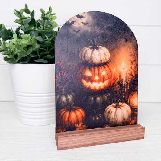 A Halloween-themed tabletop sign with an arched top and wooden base, featuring carved pumpkins, glowing candles, bats, and a dark smoky sky. The sign is styled on a white surface next to a potted green plant.