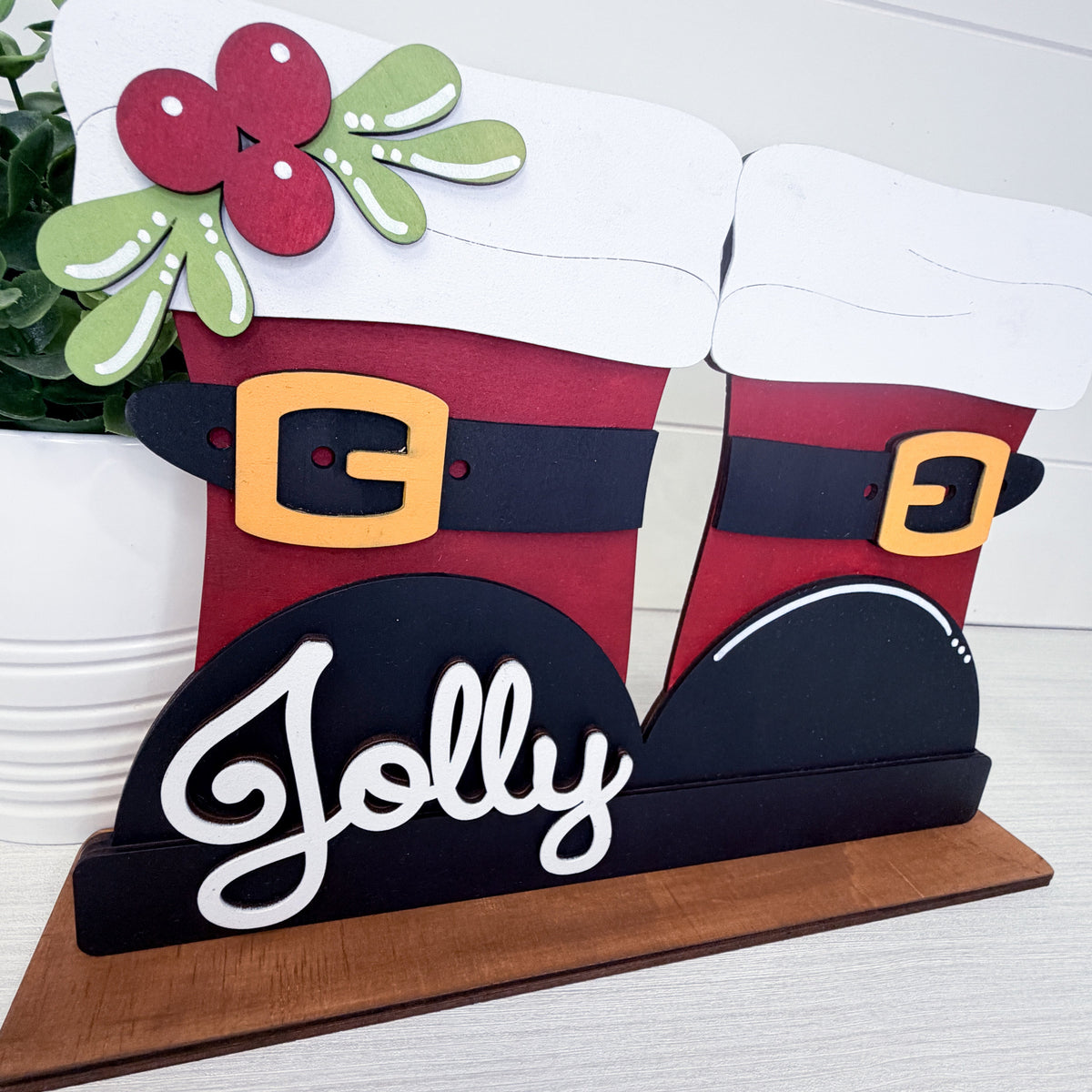 Handcrafted wooden Christmas stocking sign featuring two stockings with red bodies, black belts with gold buckles, white cuffs, a green holly leaf with a red berry at the top, and the word 'Jolly' in white script on a black base over a wooden board.