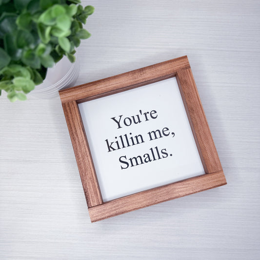 Wooden frame with a white sign inside displaying text, placed on a light surface with a plant in the corner.