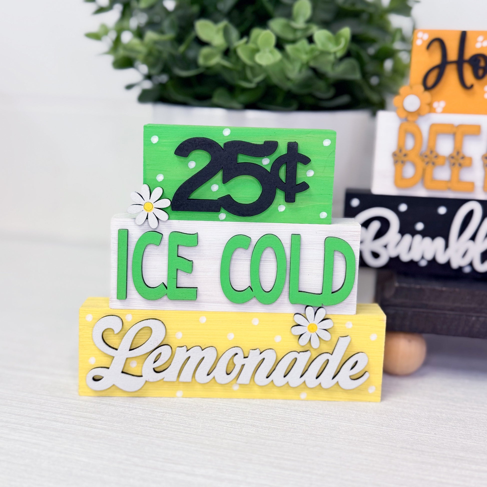 Stack of decorative signs with text '25 cents, Ice Cold Lemonade' on a white surface with a plant in the background.