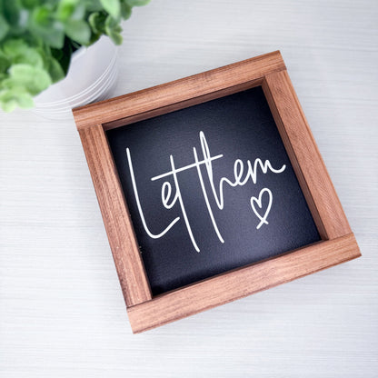 Small wooden-framed sign with 'Let them' text on a white surface with greenery.