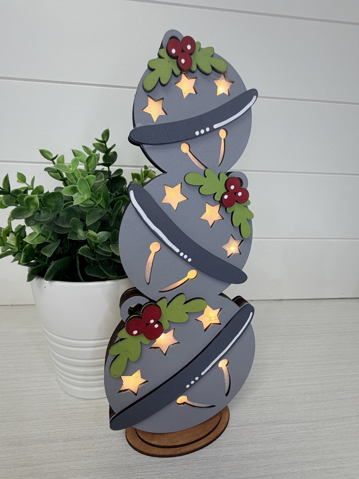 A decorative wooden Christmas tree ornament shaped like a stacked spiral of three gray Christmas tree silhouettes, adorned with gold stars, green holly leaves, red berries, and dark gray branches, displayed on a wooden stand next to a potted green plant.