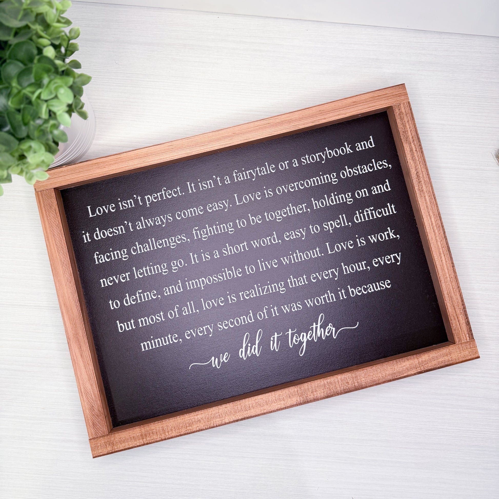 Wooden-framed plaque with a romantic quote on a white surface with a plant in the corner.