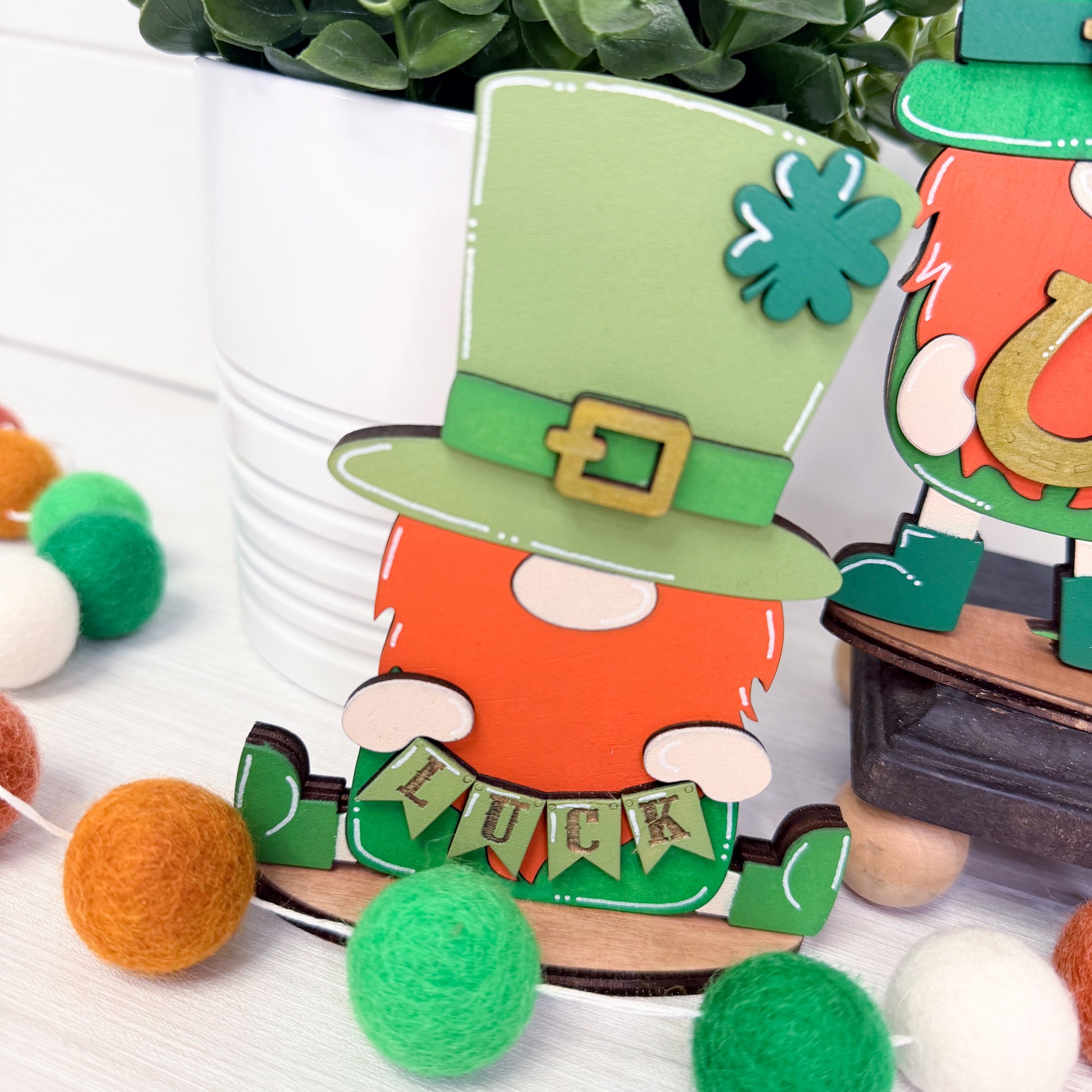Decorative wood cutout of a leprechaun with green hat and orange body, surrounded by colorful pom-poms on a white background.