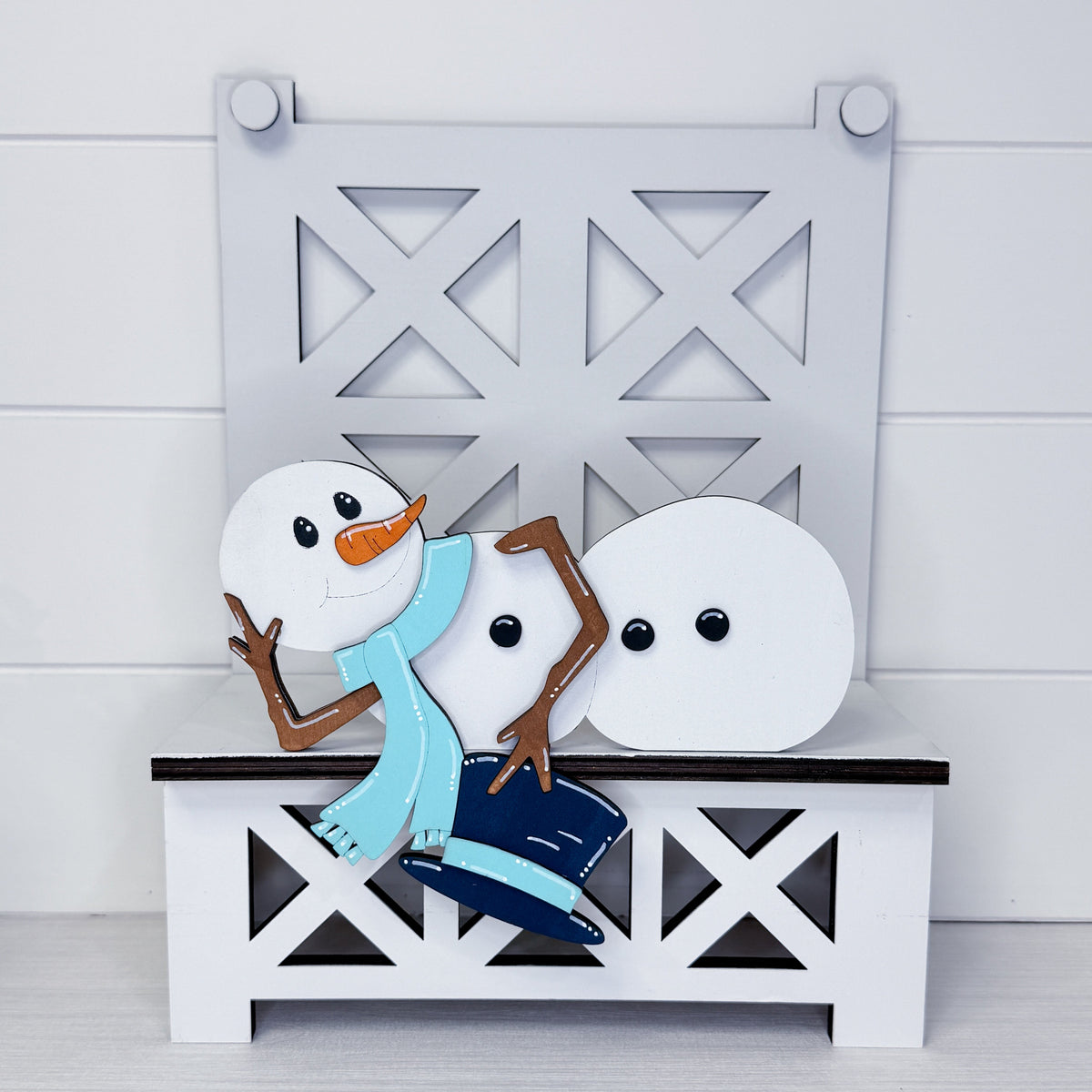 A decorative winter-themed wooden cutout set featuring three snowman figures, one wearing a light blue scarf and dark blue pants, posed on a white bench in front of a matching white lattice-patterned backdrop.