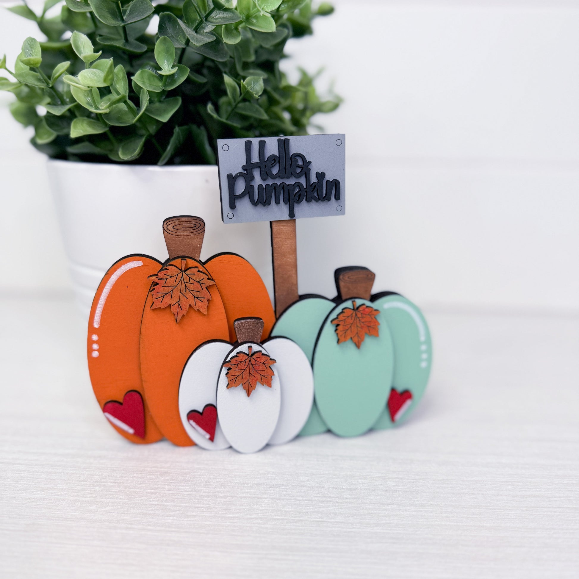 A decorative autumn-themed tabletop sign featuring three layered wooden pumpkins in orange, white, and mint green, each with a brown stem and leaf detail, plus a small sign reading 'Hello Pumpkin' on a stick, set on a light surface with a potted green plant in the background.