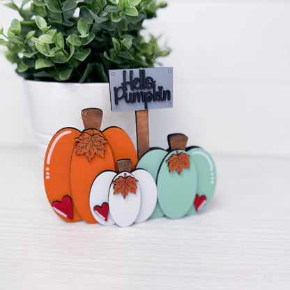 A decorative autumn-themed tabletop sign featuring three layered wooden pumpkins in orange, white, and mint green, each with a brown stem and leaf detail, plus a small sign reading 'Hello Pumpkin' on a stick, set on a light surface with a potted green plant in the background.