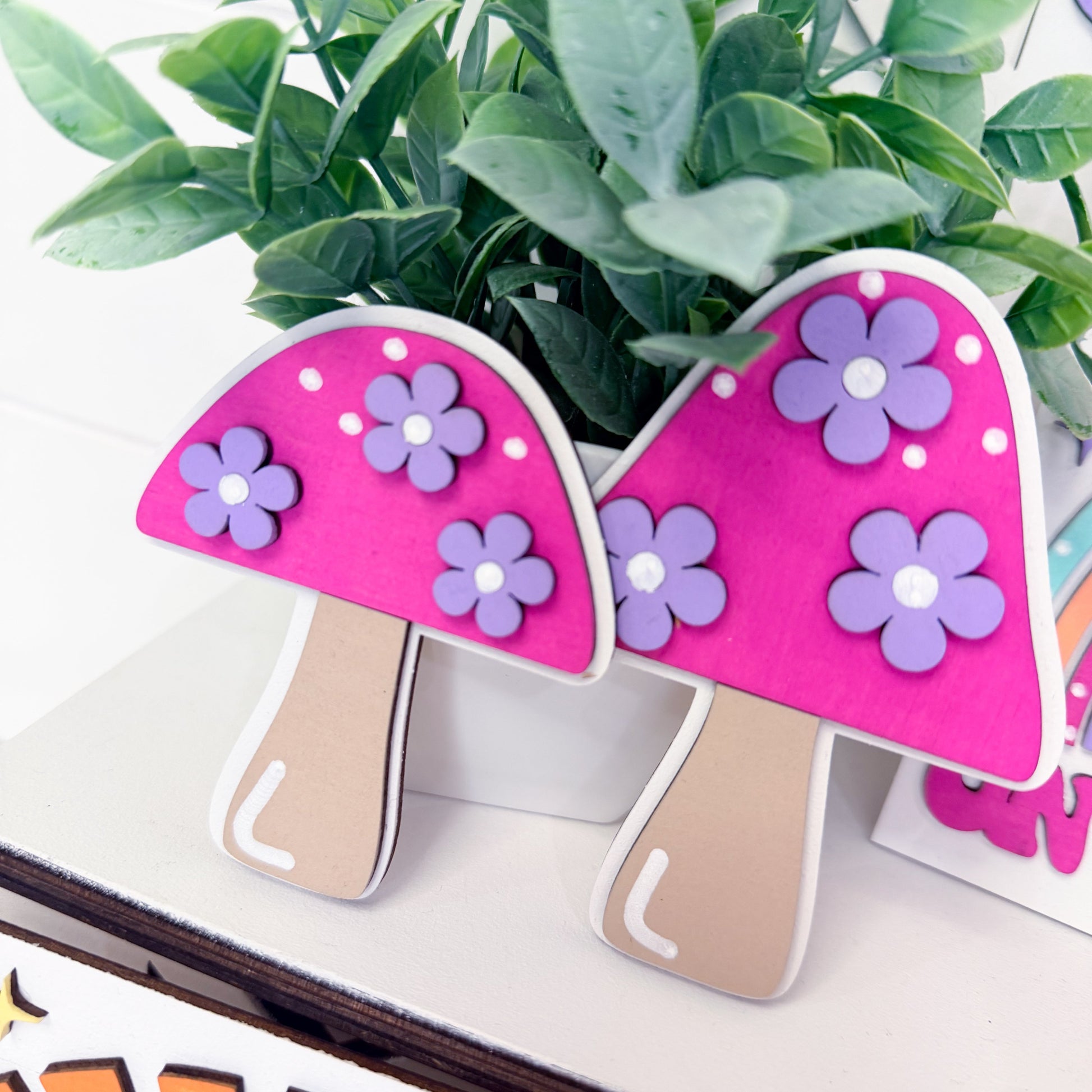 Decorative mushrooms with pink caps and purple flowers on a white surface with green leaves in the background.