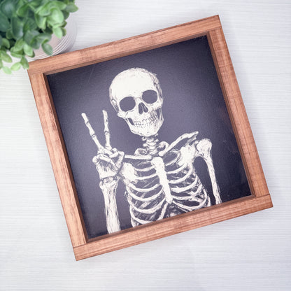 A square framed art print featuring a white skeleton drawing on a dark charcoal background, set in a light brown wooden frame and photographed from above on a white wood surface with a small green plant in the corner.