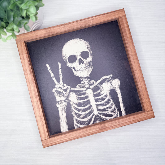 A square framed art print featuring a white skeleton drawing on a dark charcoal background, set in a light brown wooden frame and photographed from above on a white wood surface with a small green plant in the corner.