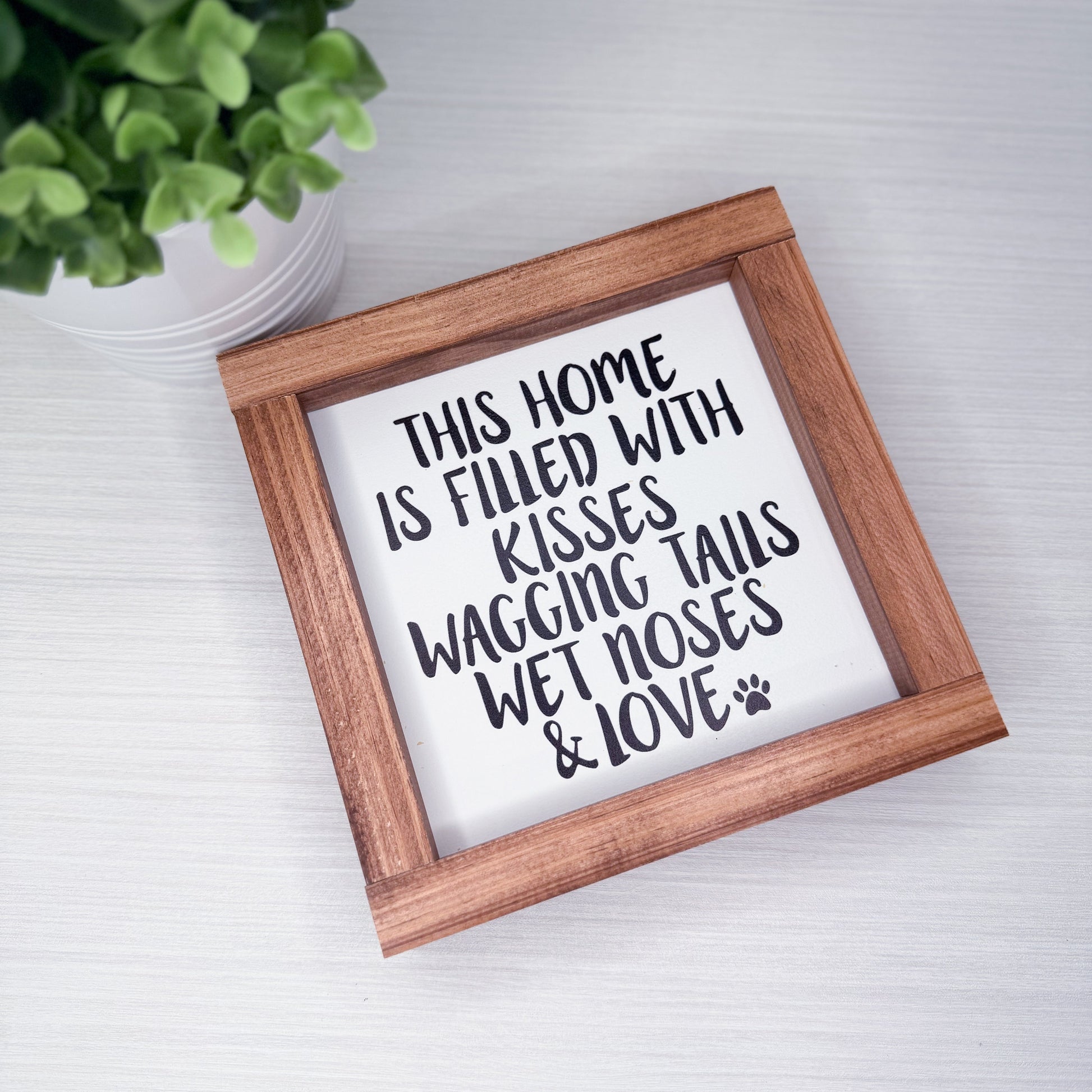Wooden-framed sign with text on a white surface with a plant in the background