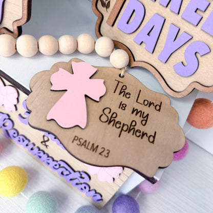 Decorative wooden tag with pink angel and 'The Lord is my Shepherd' text, surrounded by colorful beads.