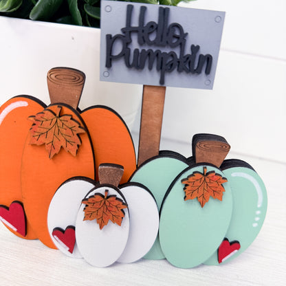 A set of autumn-themed decorative pumpkin cutouts in orange, mint green, and white, each with a brown faux-stem, a small orange leaf, and a red heart. One pumpkin features a layered 'Hello Pumpkin' sign on a wooden stick, displayed on a white wooden surface.
