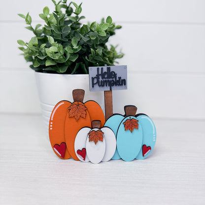 Three small decorative pumpkin ornaments in orange, white, and light blue with brown stems and leaves, arranged in front of a potted green plant on a white surface. A small sign reads “Hello Pumpkin.