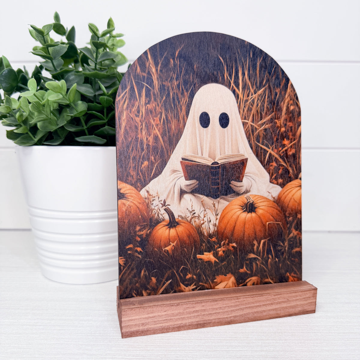 A Halloween-themed wooden plaque featuring a ghost reading a book among pumpkins, displayed on a wooden stand next to a potted green plant.