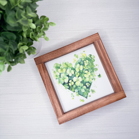 Framed artwork of a green heart on a white background with a plant in the corner.