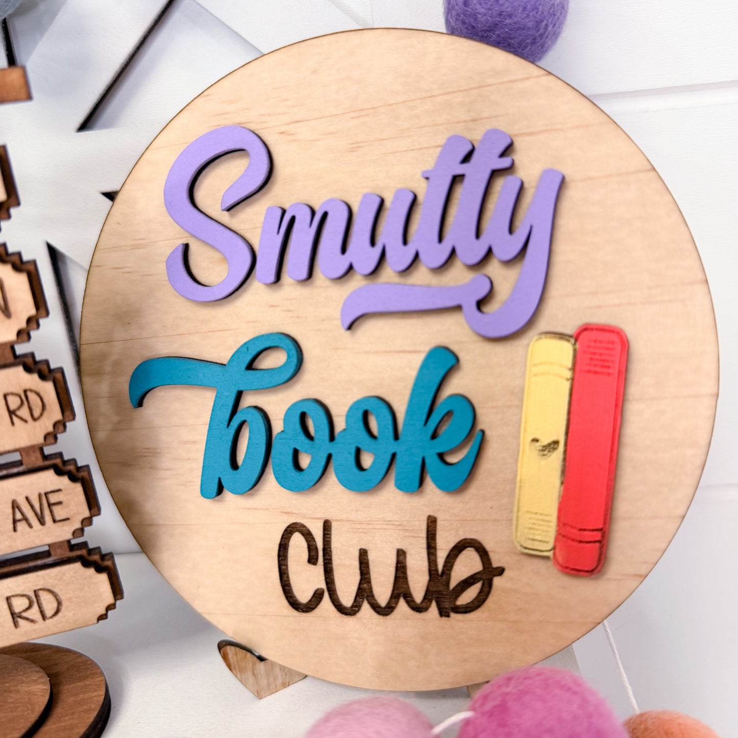 Wooden round sign with 'Smuttly Book Club' text and colorful bookends on a white background.
