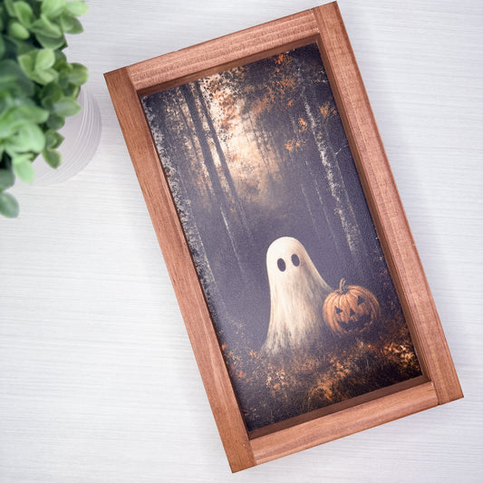 A rectangular wooden picture frame displaying a spooky Halloween illustration of a ghost and a jack-o'-lantern in a dark forest, shown on a light wood tabletop with a green plant in the corner.