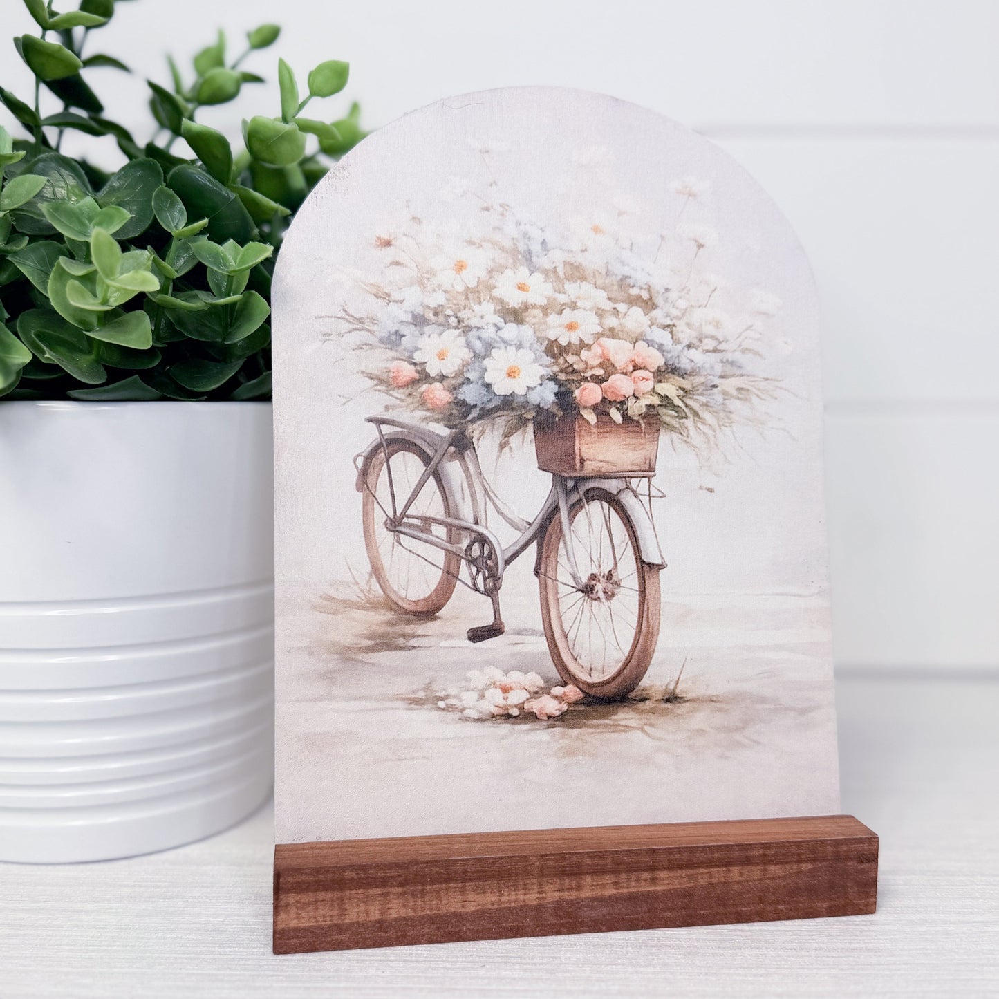 Decorative arch with bicycle and flowers design on a wooden base, next to a potted plant.