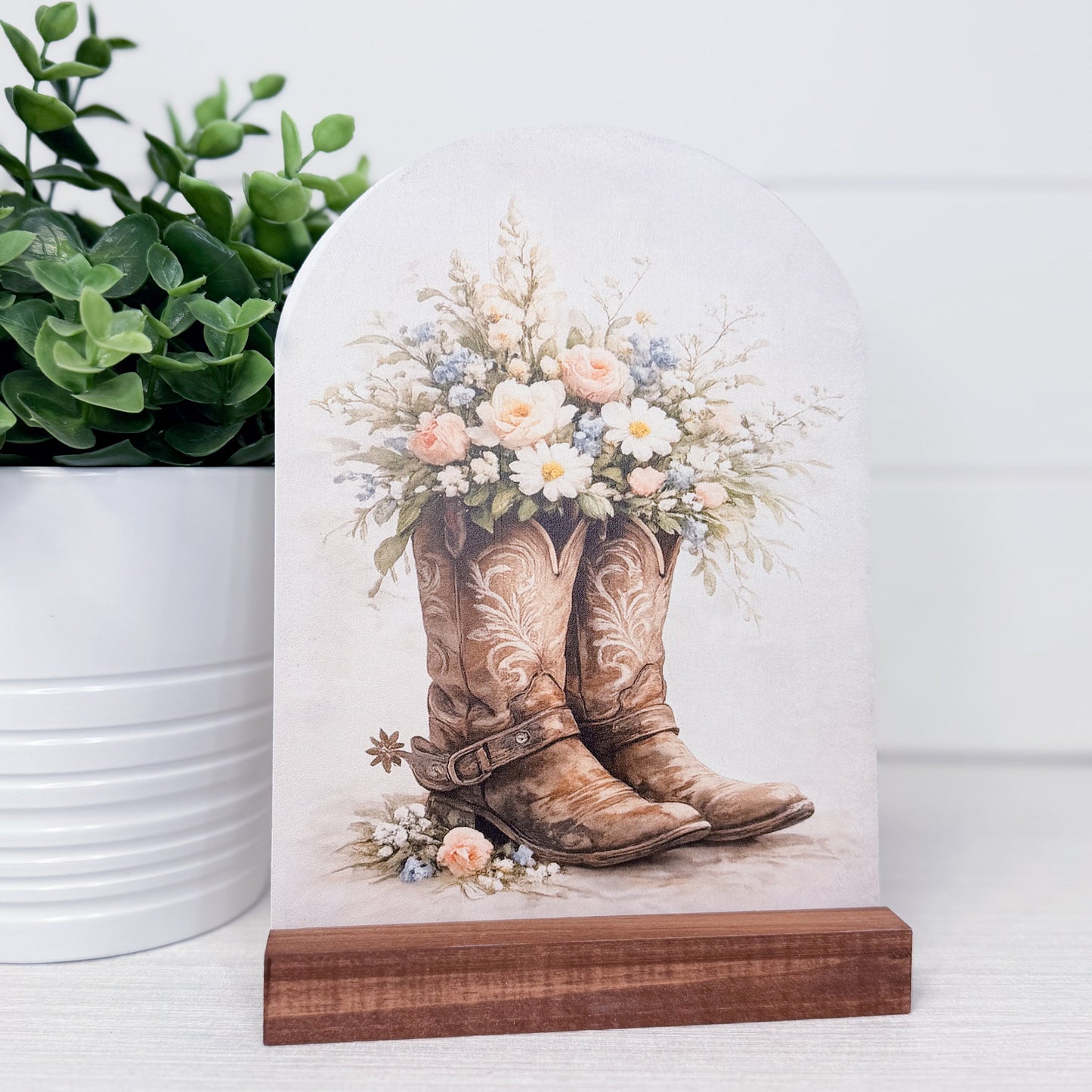 Decorative arch with cowboy boots and flowers on a wooden stand next to a potted plant.