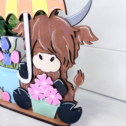 highland cow with brown hair and a colorful umbrella holding flowers.