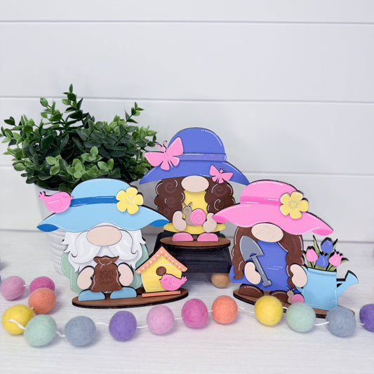 Three colorful gnome figurines with hats and accessories on a white surface with a plant and decorative balls.