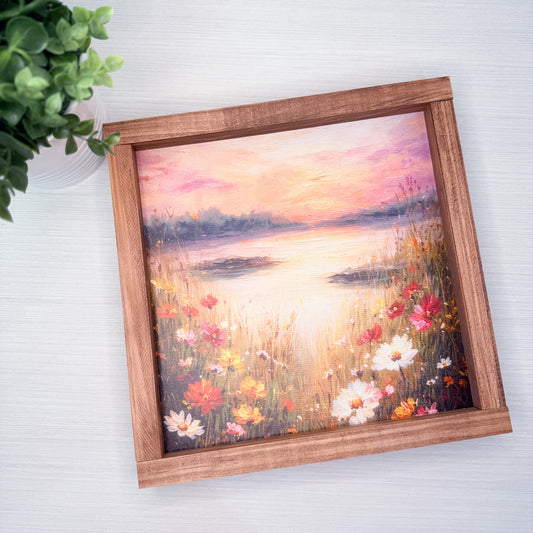 Framed painting of a sunset over water with flowers on a white surface.
