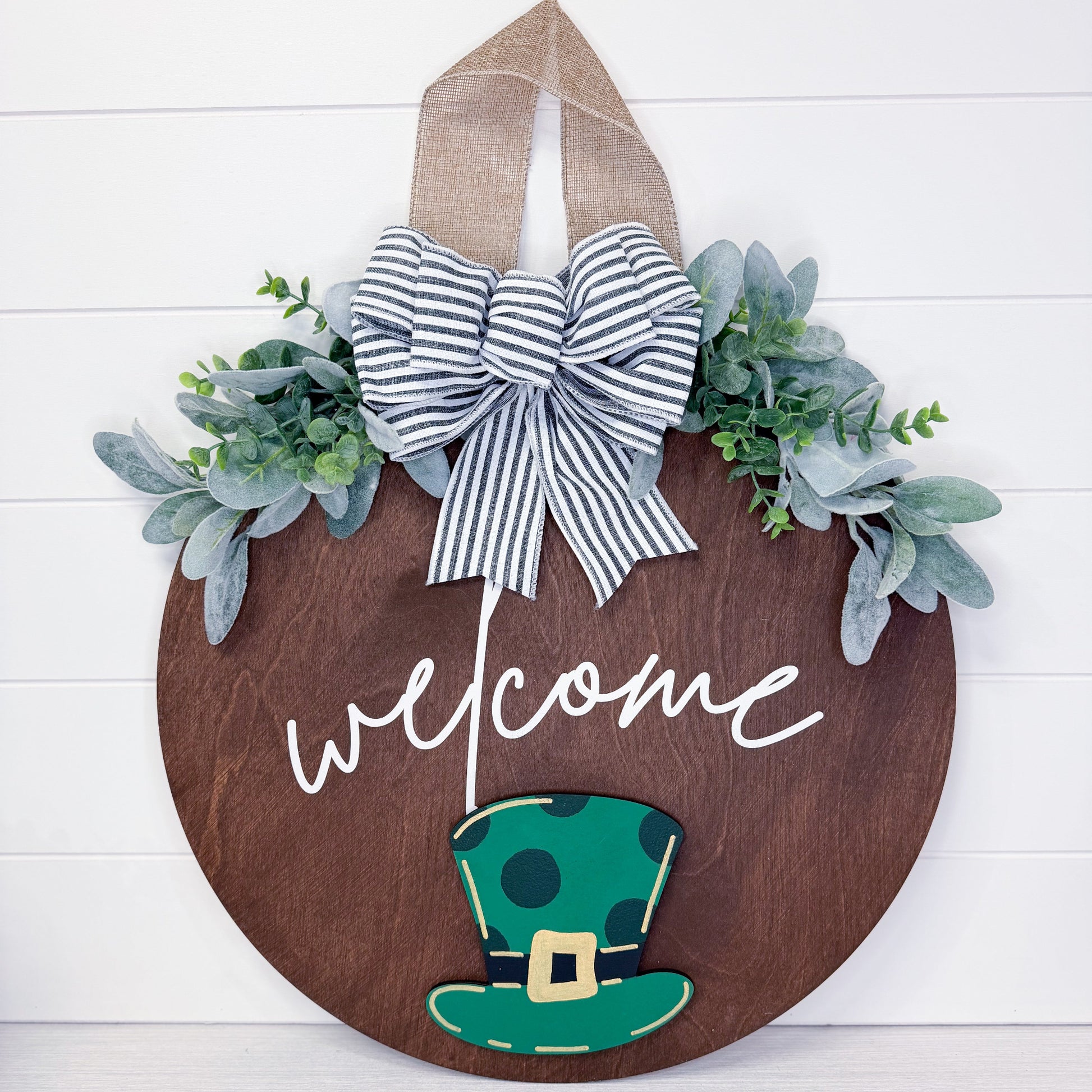 Decorative wooden door hanger with a bow, green hat, and 'welcome' text on a white background.