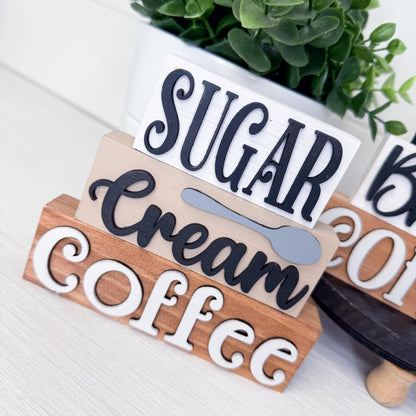 Wooden signs with 'SUGAR', 'CREAM', and 'COFFEE' text on a white surface with a plant in the background.