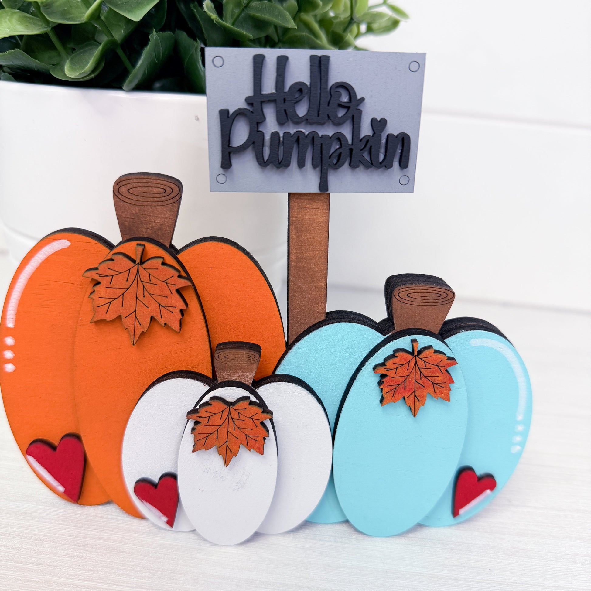 A set of decorative wooden pumpkin signs in orange, white, and light blue, each with a small red heart and an orange leaf, arranged in front of a sign that reads 'Hello Pumpkin'.