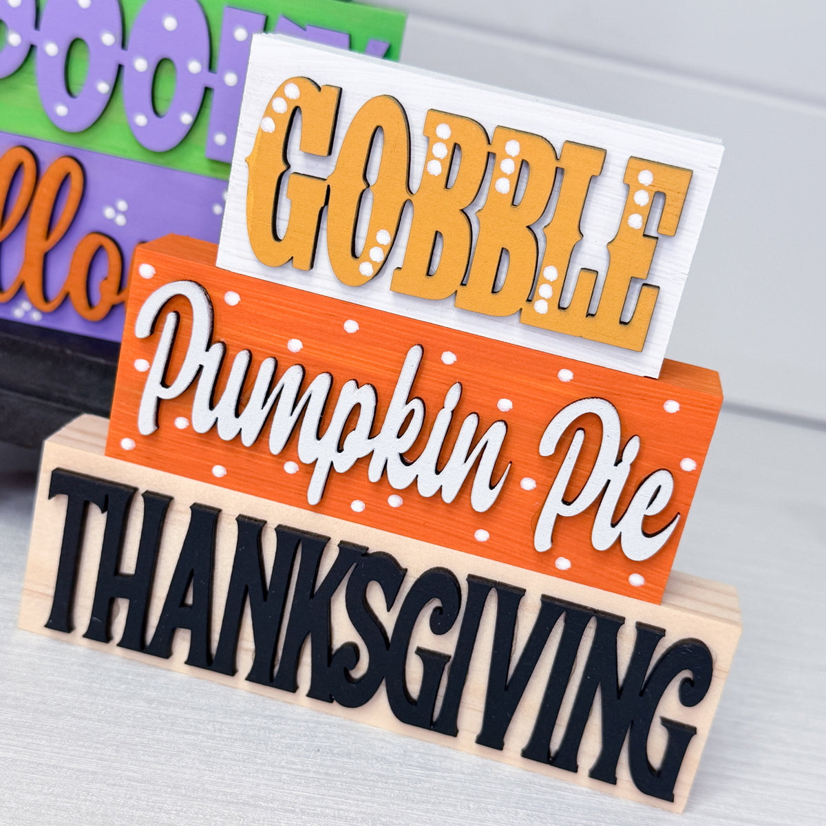 Three stacked wooden sign blocks reading 'GOBBLE', 'Pumpkin Pie', and 'THANKSGIVING' in orange, white, and black lettering, displayed on a light gray surface.