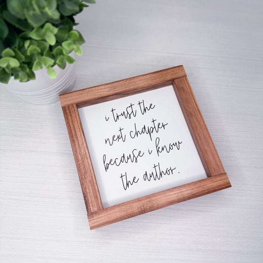 Wooden-framed sign with motivational quote on a white surface with a plant in the background