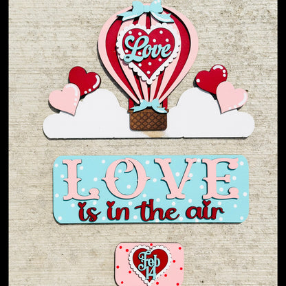 Valentine's Day-themed decor with hot air balloon, hearts, and 'Love is in the air' sign on a textured background.
