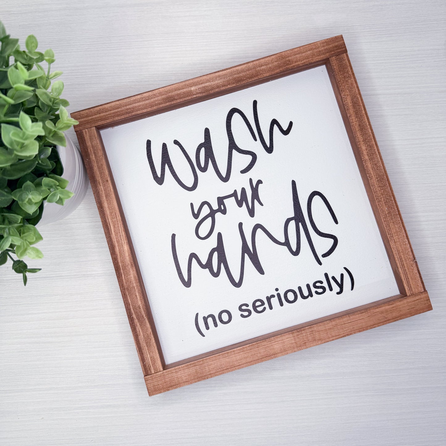 Framed sign with humorous text 'Wash your hands (no seriously)' on a light background